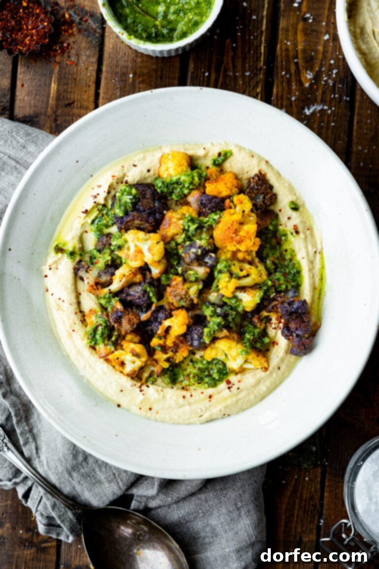 An overhead view of a serving plate filled with creamy hummus, generously topped with an array of vibrant, roasted tricolor cauliflower florets, offering a visually appealing and healthy meal option.