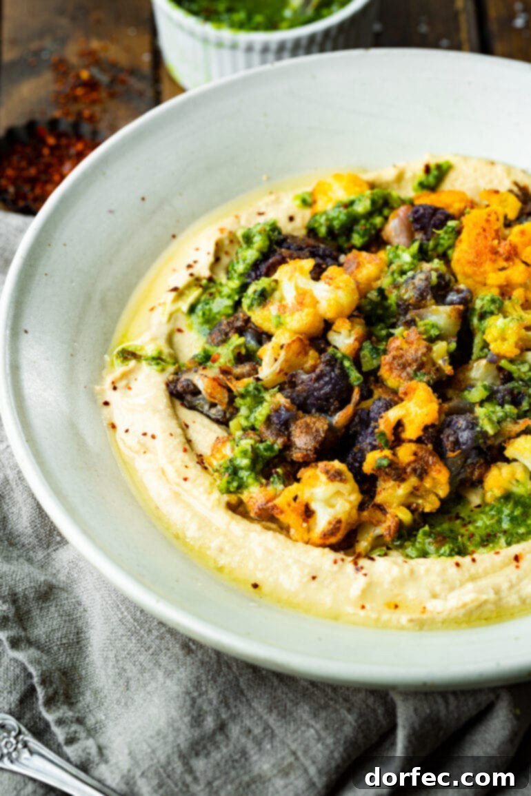 A beautifully presented serving bowl filled with creamy hummus, generously topped with roasted tricolor cauliflower florets and artfully drizzled with fresh pesto sauce, creating an appealing and healthy dish.
