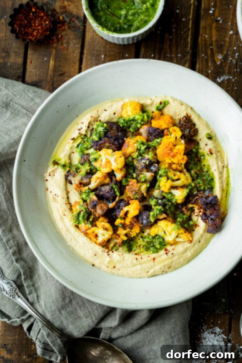 A high-angle shot of a serving bowl featuring creamy hummus topped generously with crispy, golden roasted cauliflower florets, beautifully drizzled with vibrant pesto sauce, ready to be served.