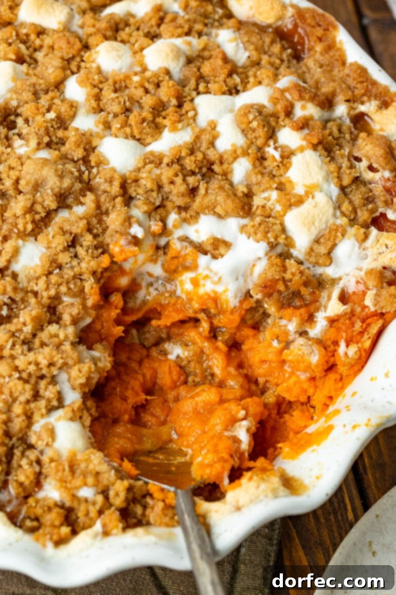a photo of a round baking dish full of sweet potato casserole topped with gooey marshmallows and a brown sugar streusel topping with a serving being scooped out of the dish