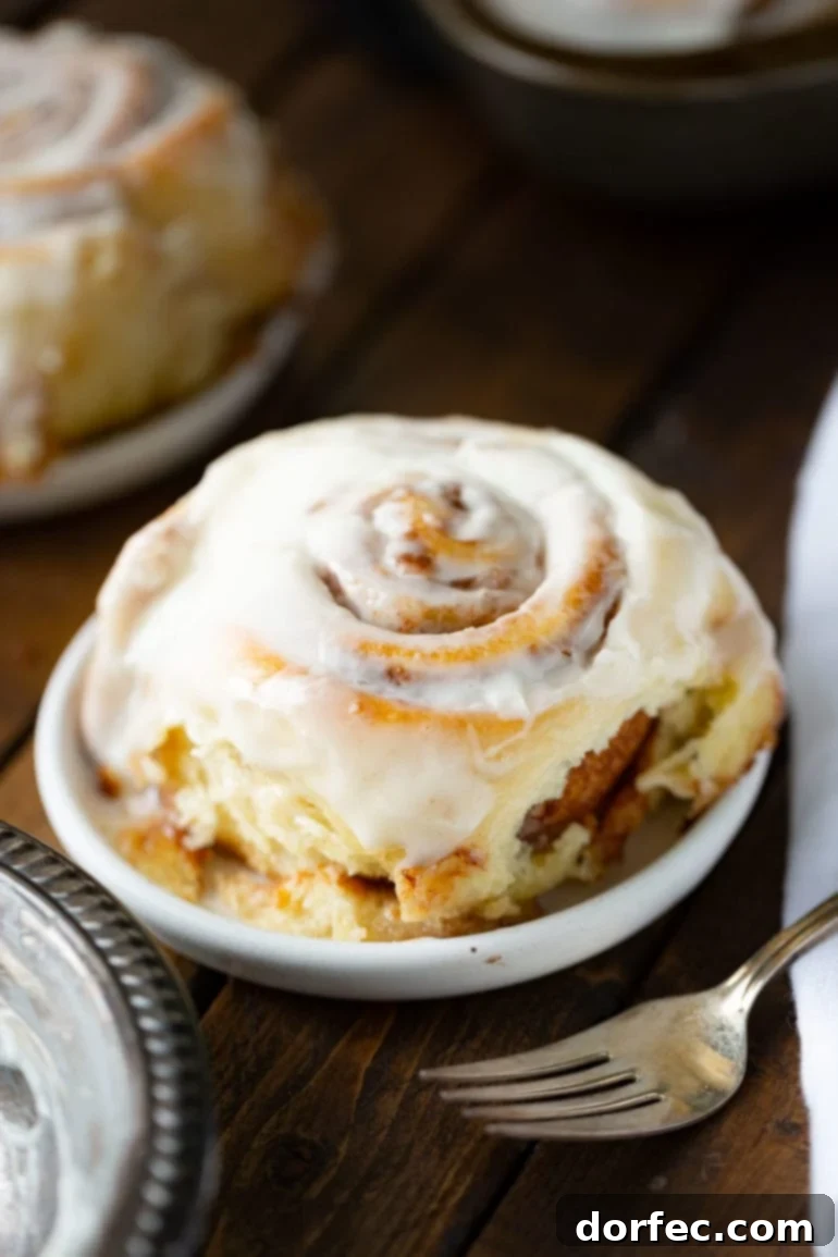 a photo of a single cinnamon roll topped with a creamy white frosting sitting on a small dessert plate with a silver fork in the foreground.