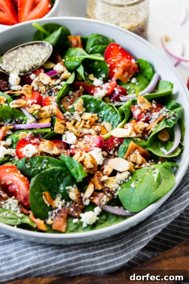 a photo of a large white serving bowl full of a spinach salad topped with sliced strawberries, sliced red onions, candied slivered almonds, feta cheese, bacon crumbles and homeade poppy seed dressing.