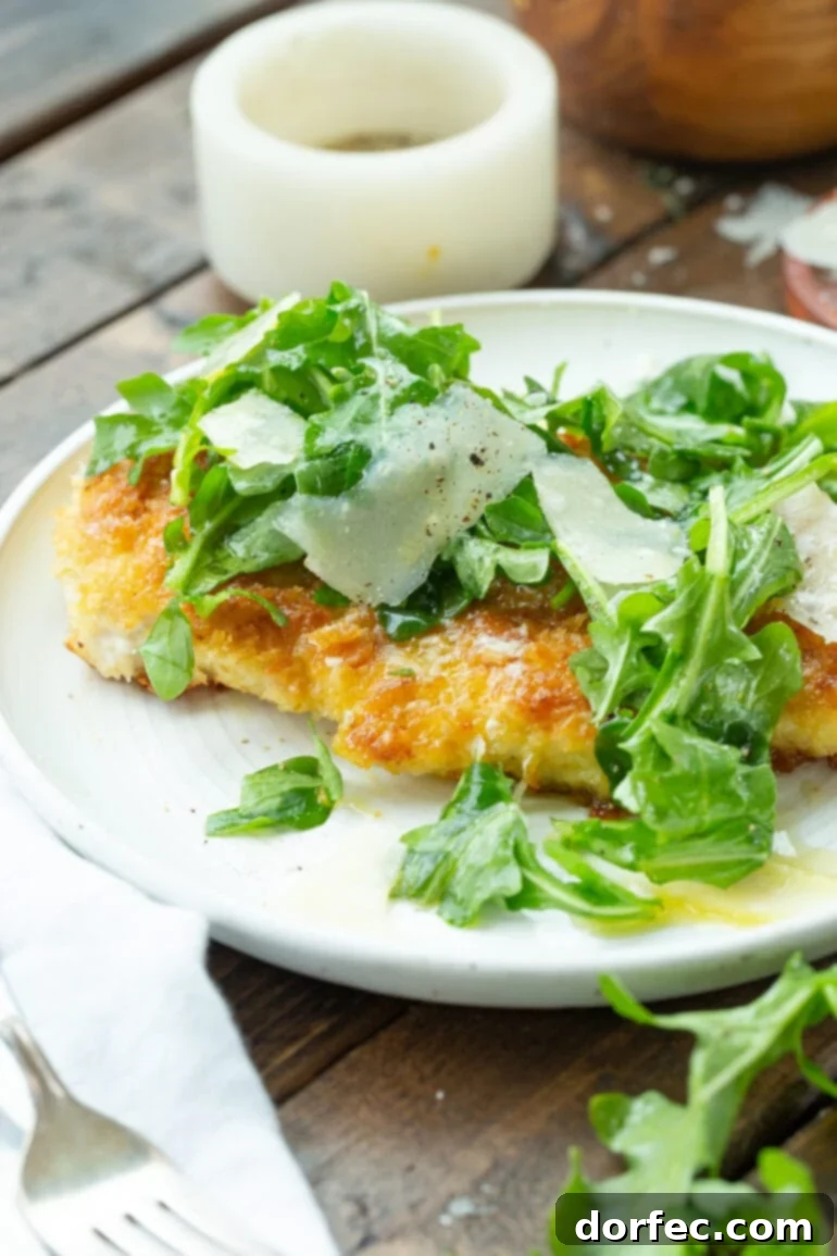 a photo of a golden lightly breaded chicken breast sitting on a white dinner plate topped with salad greens and shaved parmesan.