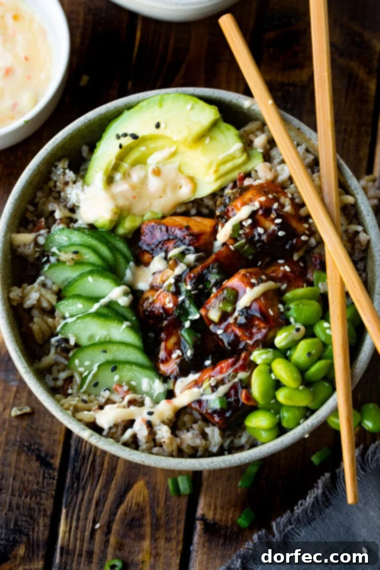 a photo of a bowl full of rice topped with teriyaki salmon chunks, edamame beans, sliced of cucumber and avocado topped with sesame seeds. there is a pair of wooden chopsticks sitting crossed on the side of the bowl.
