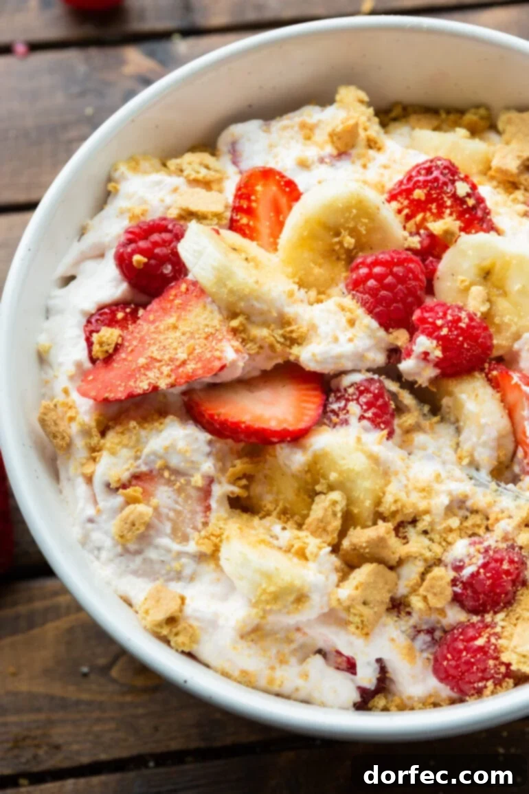 Dreamy Fluffy Cheesecake Fruit Salad 3 A close-up shot of a creamy cheesecake fruit salad, featuring colorful slices of banana and strawberries, plump whole raspberries, and scattered crushed graham cracker pieces, inviting to eat.