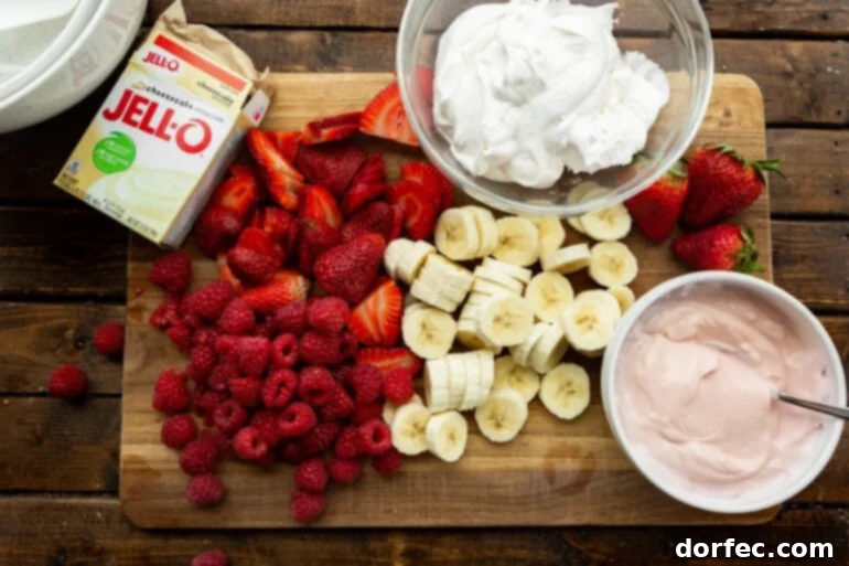Dreamy Fluffy Cheesecake Fruit Salad 4 A flat lay photograph showcasing all the fresh ingredients laid out on a wooden cutting board: neatly sliced bananas, bright strawberries, whole raspberries, alongside bowls of strawberry yogurt, a box of cheesecake pudding mix, and a container of whipped cream.