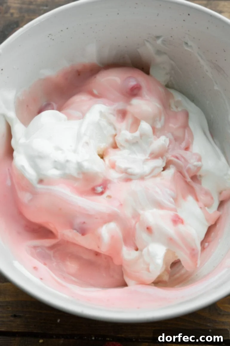 Dreamy Fluffy Cheesecake Fruit Salad 5 A close-up of a cheesecake pudding mix being stirred vigorously with strawberry yogurt and Cool Whip in a large bowl, creating a smooth and creamy pink base for the fruit salad.