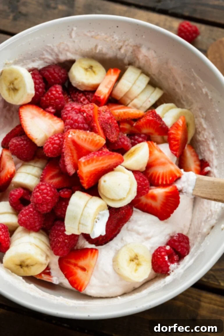 Dreamy Fluffy Cheesecake Fruit Salad 6 Sliced strawberries, bananas, and whole raspberries being gently folded into a light pink creamy yogurt mixture in a large bowl, demonstrating the careful combination of ingredients.