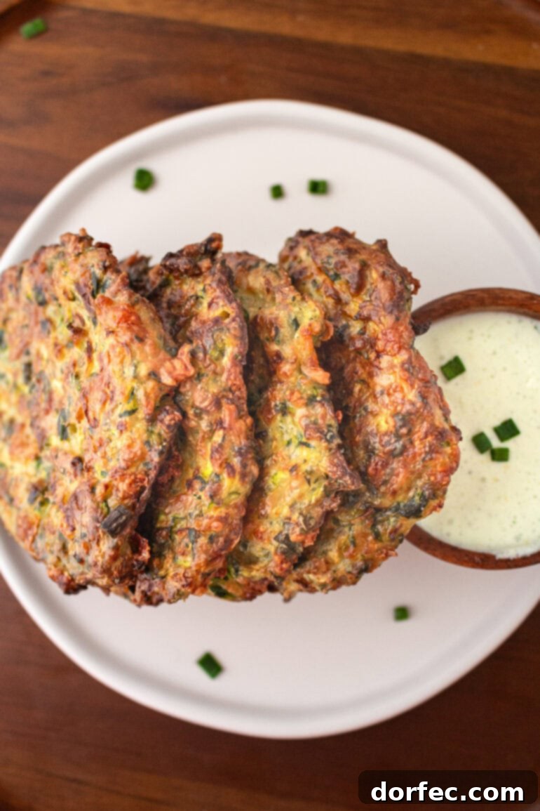 Crispy Parmesan Basil Zucchini Fritters 11 A delicious stack of four crispy zucchini fritters, showcasing their golden-brown exterior, leaning against a small cup of creamy dipping sauce.