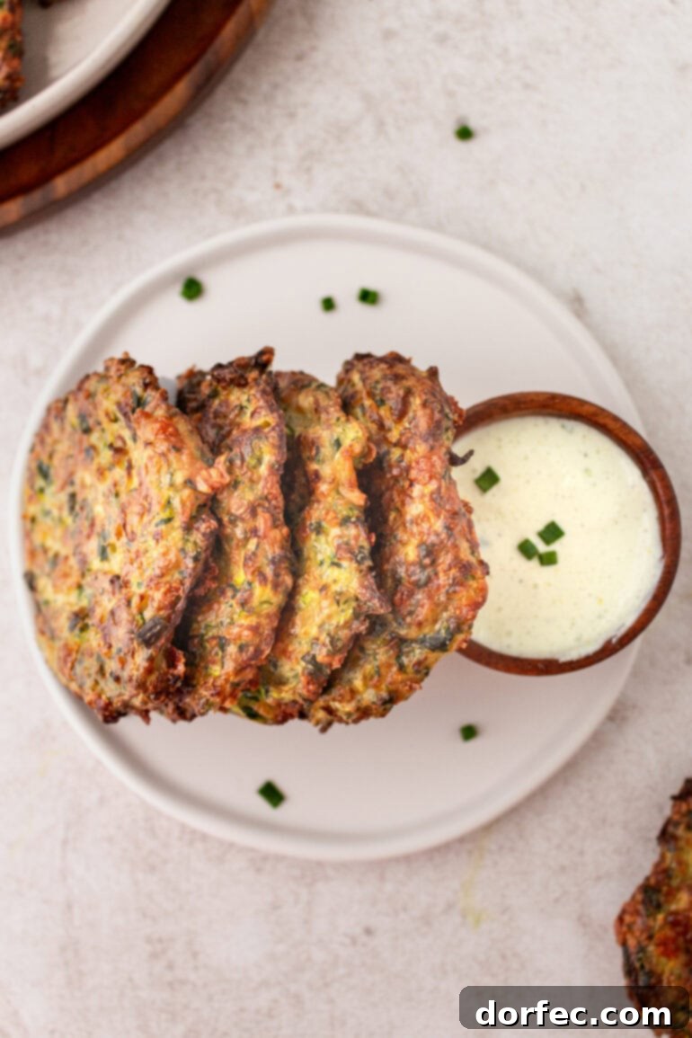 Crispy Parmesan Basil Zucchini Fritters 4 A close-up shot of golden, crispy zucchini fritters, artfully arranged and leaning against a small cup of creamy dipping sauce on a rustic plate.