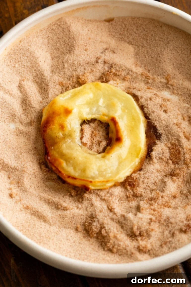 A single golden-battered fried apple ring, just out of the air fryer, being coated in a bowl of crunchy cinnamon sugar.