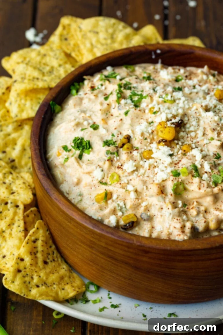 A large wooden bowl filled with vibrant Mexican Street Corn Dip, surrounded by scattered tortilla chips, ready for dipping.