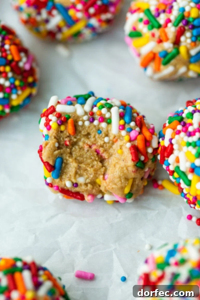 A close-up photo of several delicious no-bake birthday cake protein balls, adorned with colorful rainbow sprinkles, with one ball having a bite taken out of it to show its soft interior.