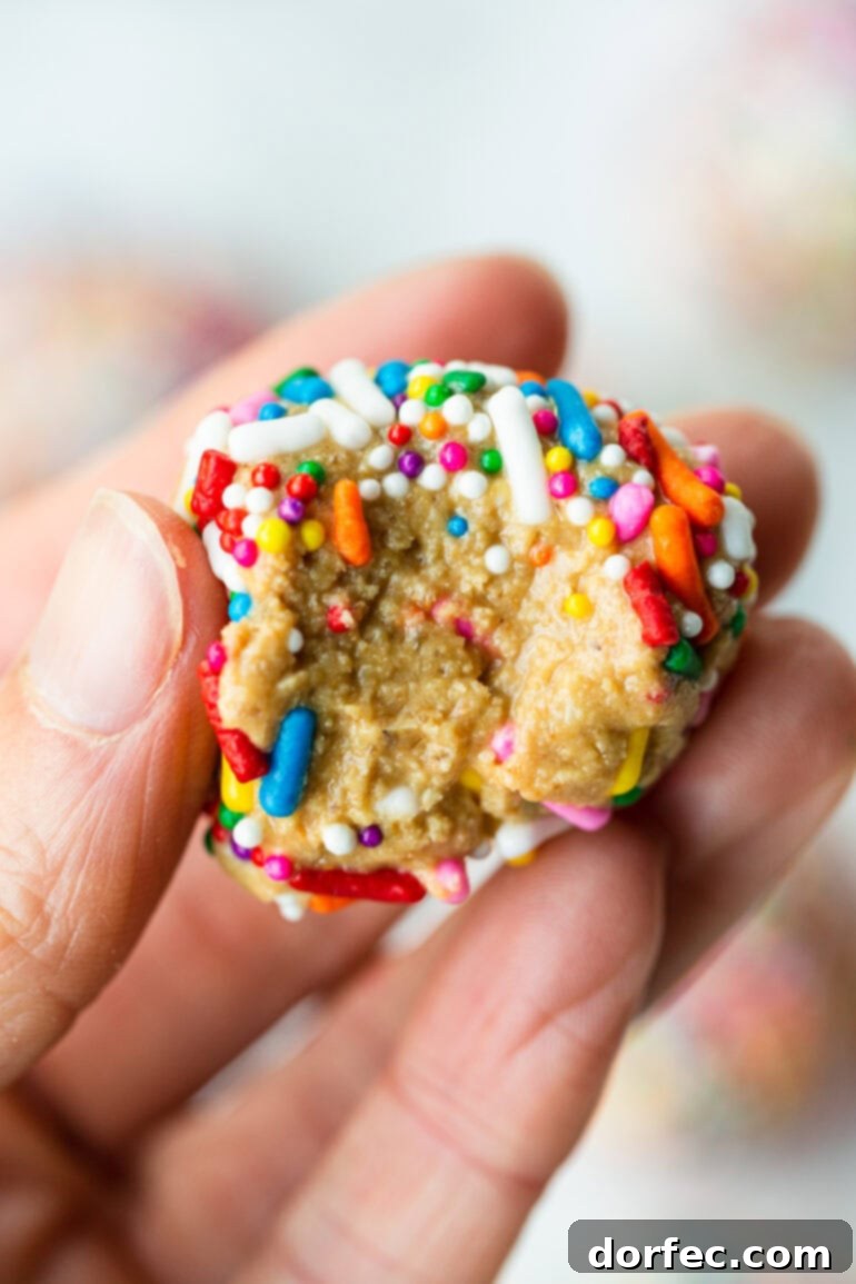 A close-up photo of someone holding a single birthday cake protein ball, covered in rainbow sprinkles, with a small bite taken out of it, showcasing the soft interior.