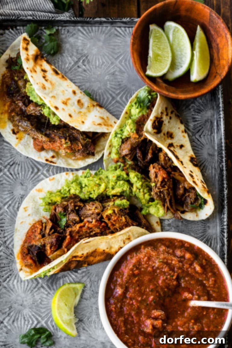 A tempting visual of several shredded beef tacos arranged on a baking sheet, accompanied by a small bowl of vibrant salsa and fresh lime wedges, ready for a delicious meal.