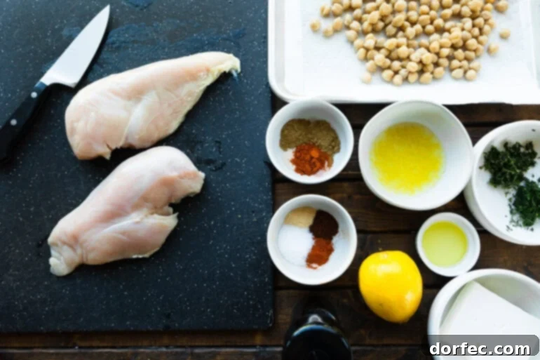 A top-down view of fresh ingredients laid out, including raw chicken breasts, a block of feta, a lemon, a can of chickpeas, and various spices, ready for cooking.