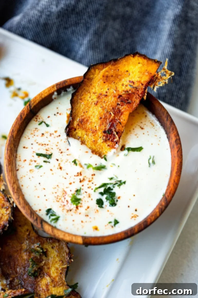 A close-up photo of a single piece of roasted acorn squash, golden-brown with crispy cheese edges, partially dipped into a small white bowl of creamy sauce.