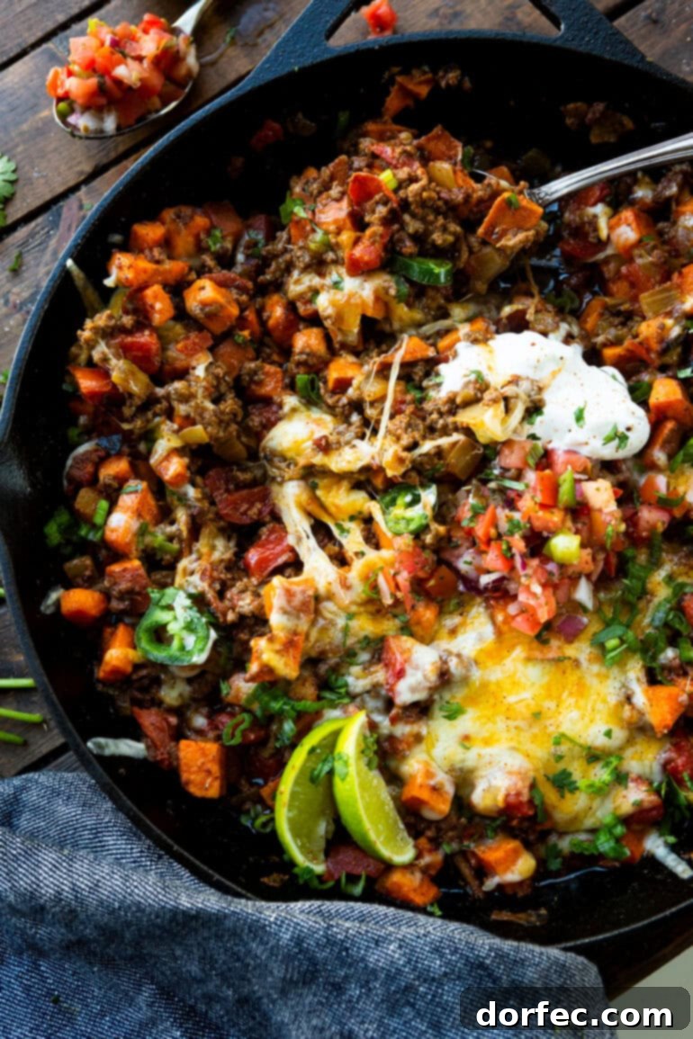 A beautifully plated cast iron skillet of cubed sweet potatoes and taco ground beef, generously topped with sour cream, fresh lime wedges, finely diced purple onions, and vibrant sliced jalapeños.