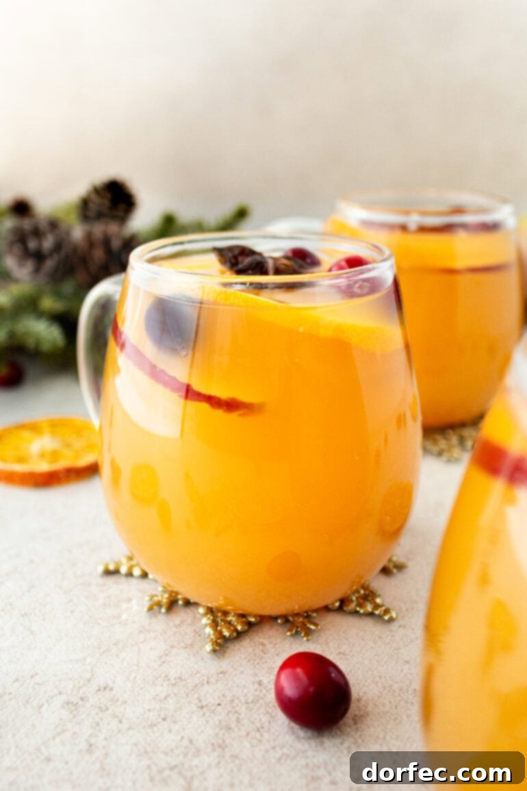A photo taken from the side of a clear glass mug full of bright orange homemade Wassail, showcasing its vibrant color and inviting warmth. This delightful Wassail recipe features a harmonious blend of fruit juices and spices, making it a standout holiday drink.