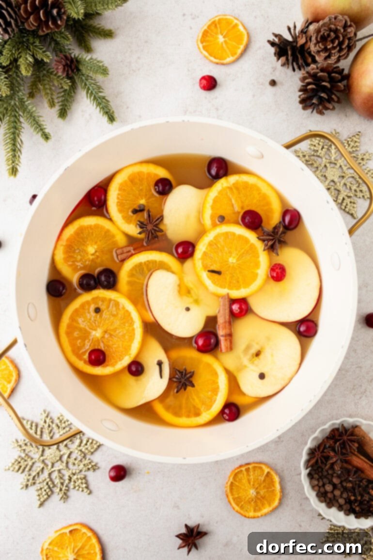 A large white pot overflowing with homemade Wassail, richly garnished with sliced apples, orange wheels, whole cranberries, and fragrant cinnamon sticks. This visually appealing and delicious holiday drink recipe is perfect for serving at festive gatherings.
