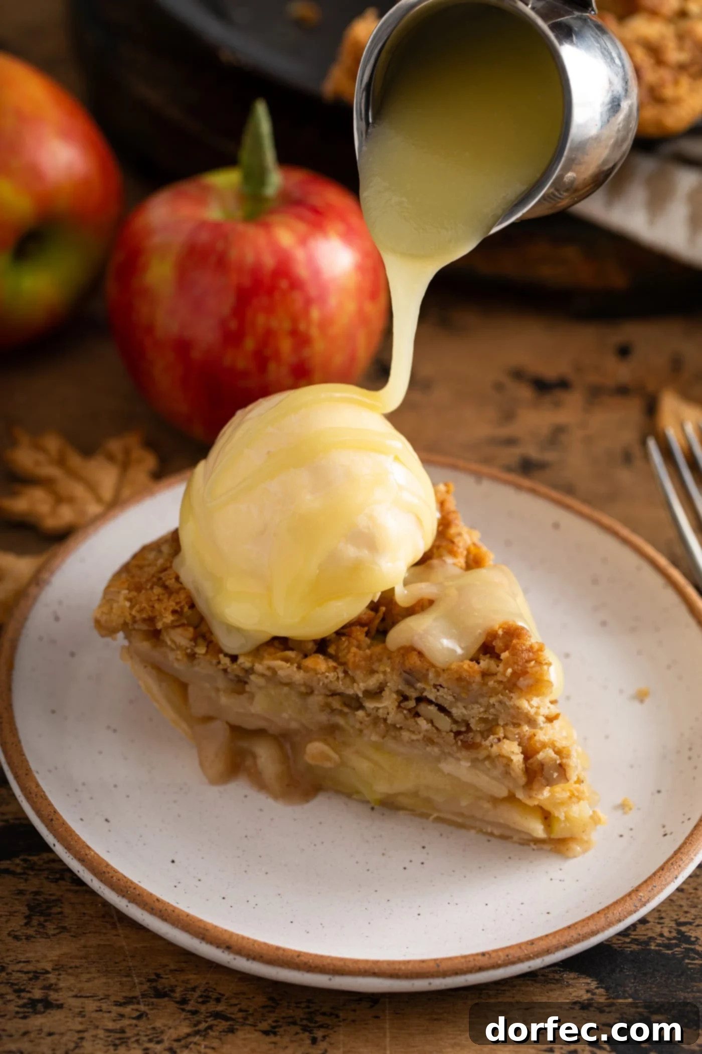 A slice of Dutch apple pie with a scoop of vanilla ice cream on top, draped generously with luscious white chocolate butter sauce. A vibrant red apple rests in the soft-focus background.