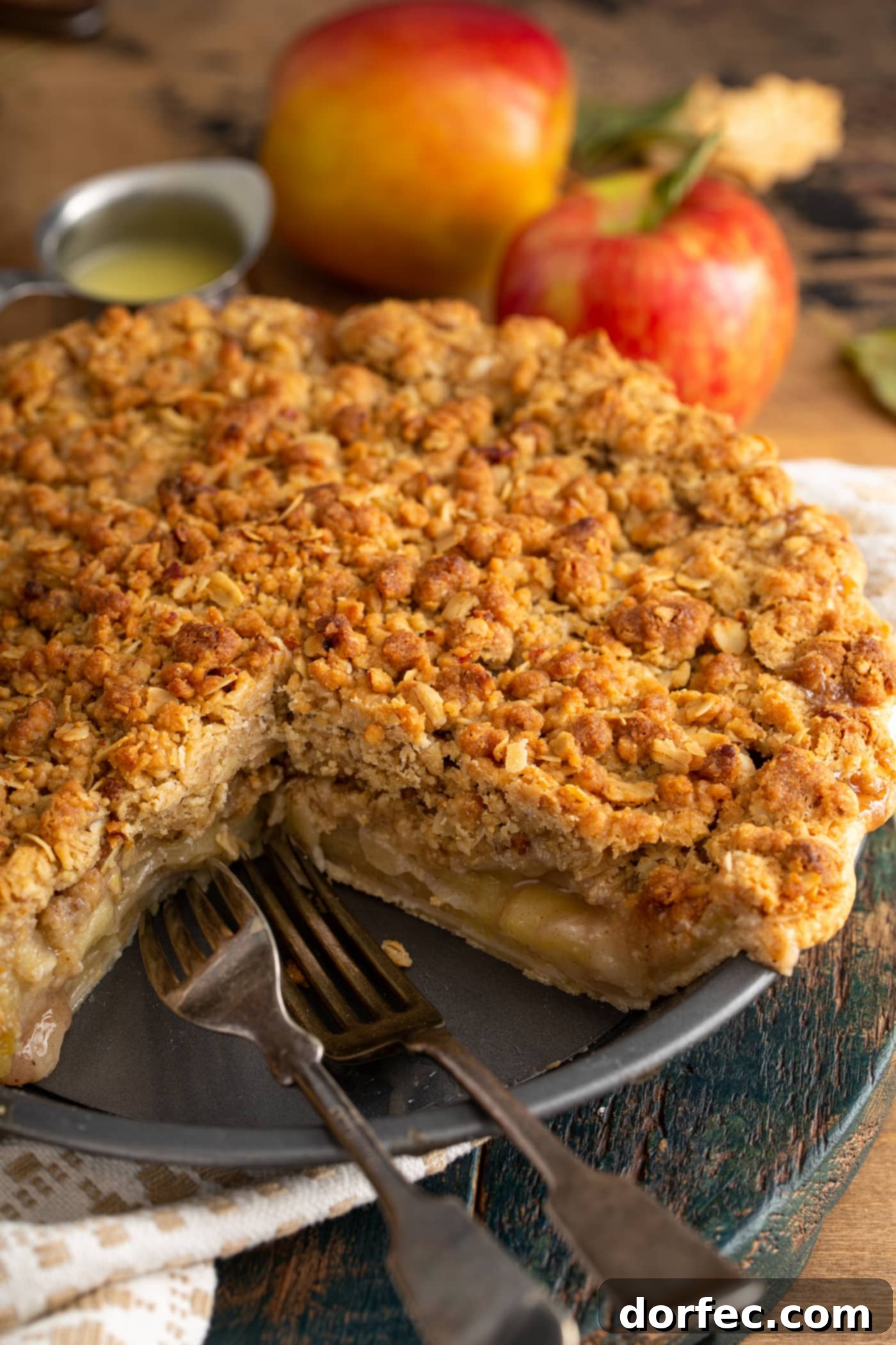 A beautifully baked Dutch apple pie with a slice removed, revealing the tender, sliced apples inside and the crunchy brown crumble on top. A couple of fresh apples are in the background, hinting at the pie's key ingredient.