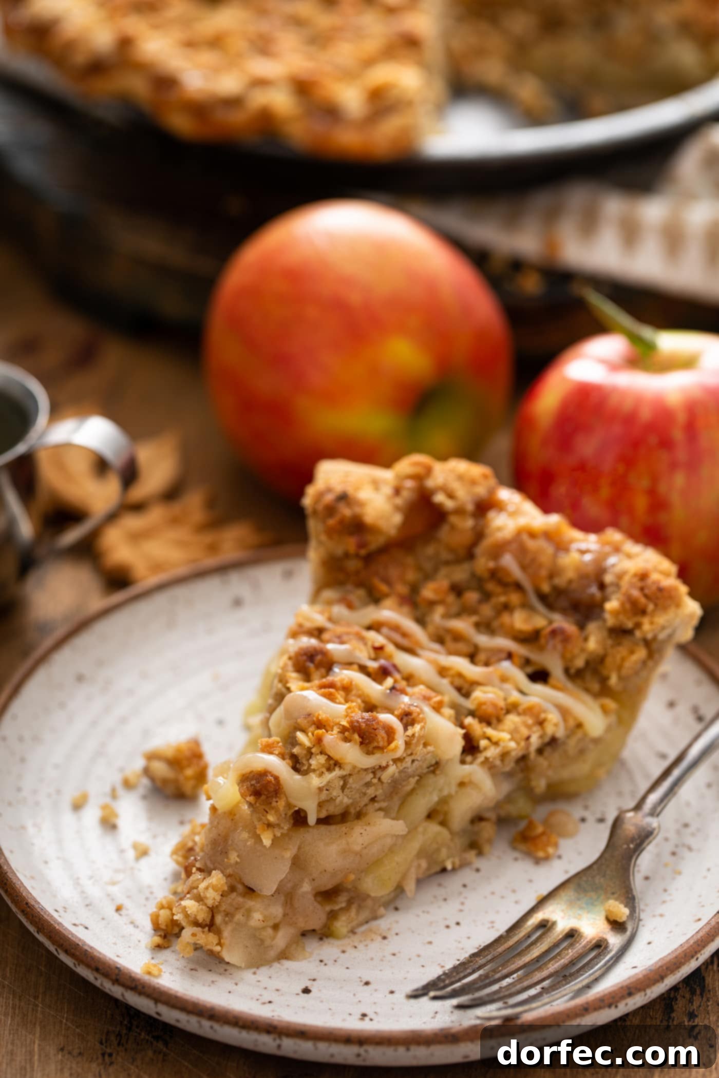 A close-up of a rustic slice of Dutch apple pie, delicately drizzled with white chocolate butter sauce. Two fresh apples provide a natural backdrop, emphasizing the pie's wholesome ingredients.