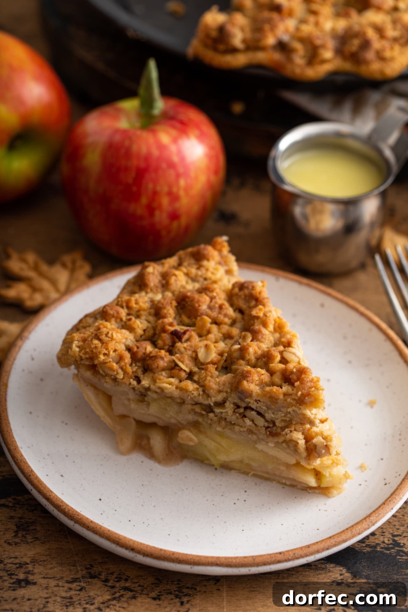 A perfect slice of Dutch Apple Pie, showcasing layers of tender apples beneath a thick, golden-brown crumble topping. A fresh apple and a jar of sauce are subtly visible in the background.