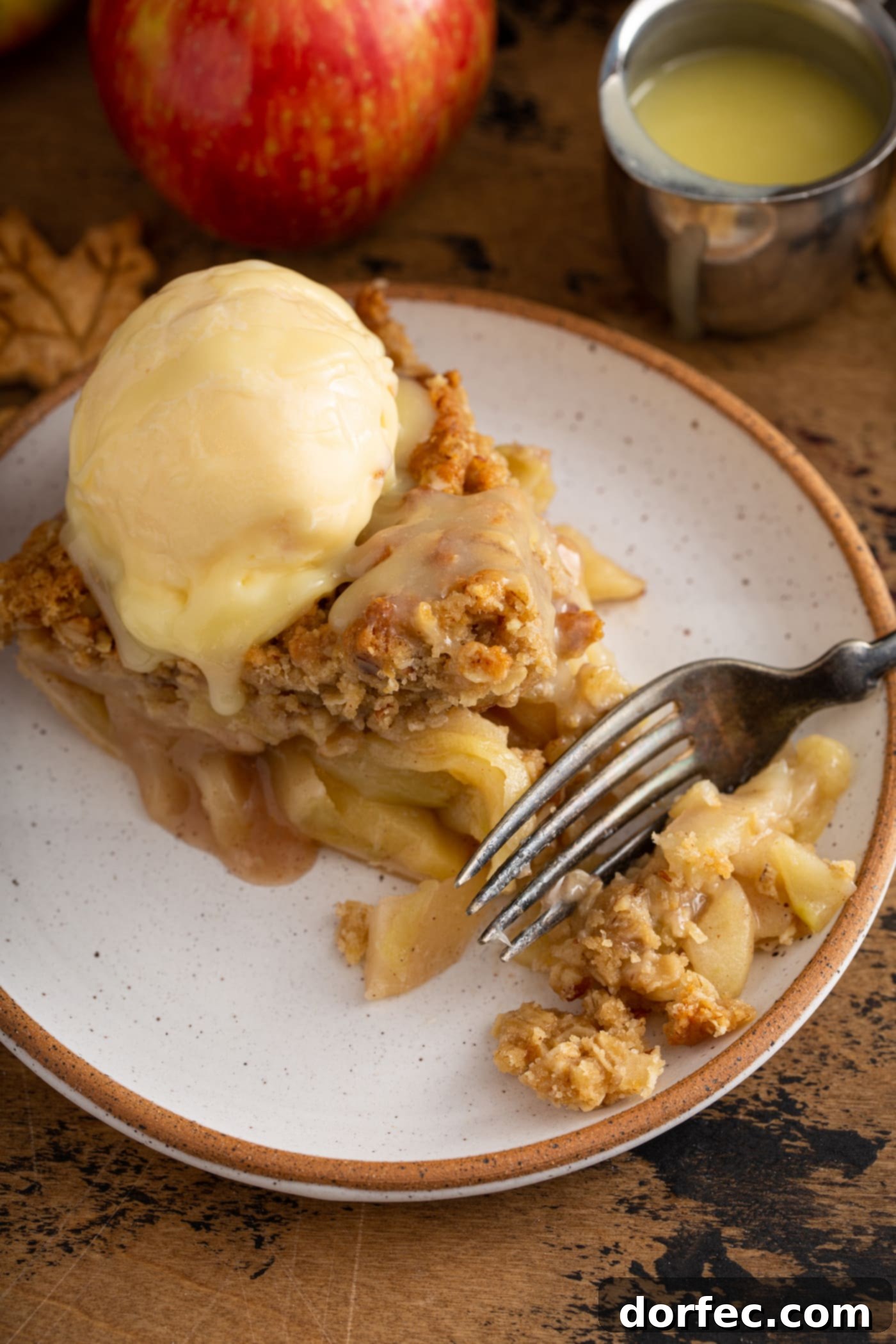 A delectable slice of Dutch apple pie served on a white dessert plate, adorned with a generous scoop of vanilla ice cream and drizzled with a rich white chocolate butter sauce. A fork rests beside the plate, inviting a taste.