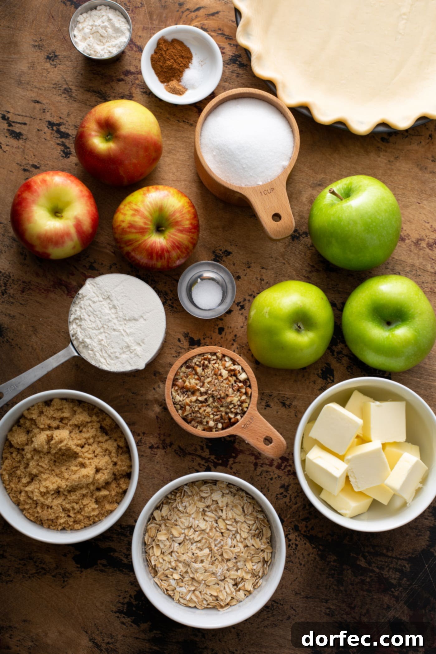 A vibrant flat lay image showcasing the fresh ingredients for Dutch Apple Pie, including crisp green and red apples, rolled oats, butter, granulated sugar, brown sugar, and an assortment of aromatic spices.