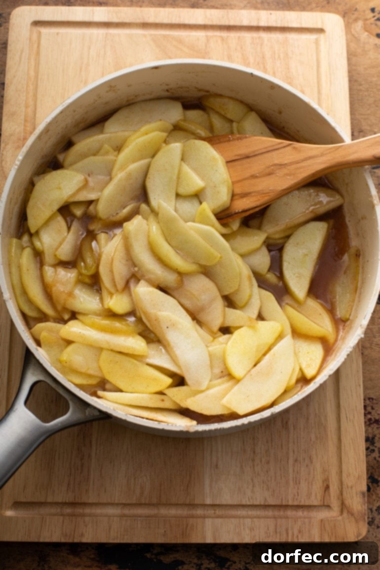 A saucepan filled with glistening sliced apples being stirred gently into melted butter and sugar, just beginning to soften for the pie filling.
