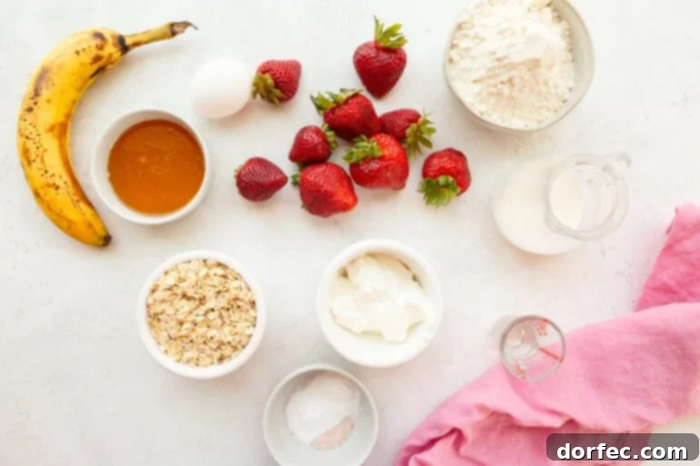 An assortment of fresh ingredients laid out on a table, including strawberries, oats, flour, Greek yogurt, honey, eggs, and a mashed banana, ready for baking healthy strawberry oat muffins.