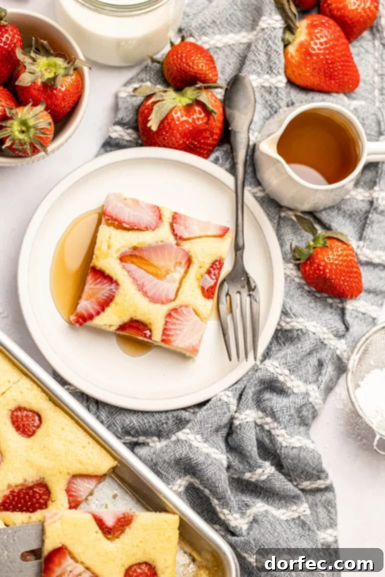 One Pan Strawberry Pancakes 14 A single serving of strawberry sheet pan pancakes on a small white plate with a bottle of syrup on the side, and the full sheet pan of baked pancakes in the background.