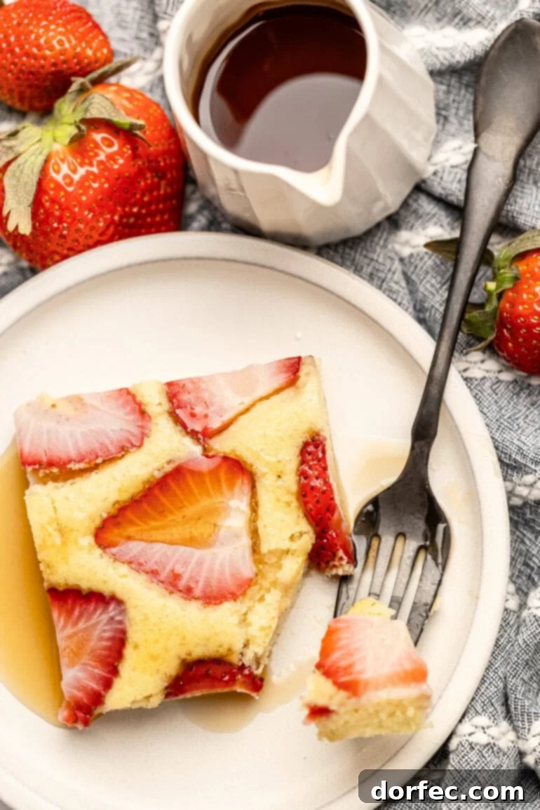 One Pan Strawberry Pancakes 3 A single slice of fluffy strawberry sheet pan pancake on a white plate, with a fork cutting a piece and a bottle of maple syrup next to it.