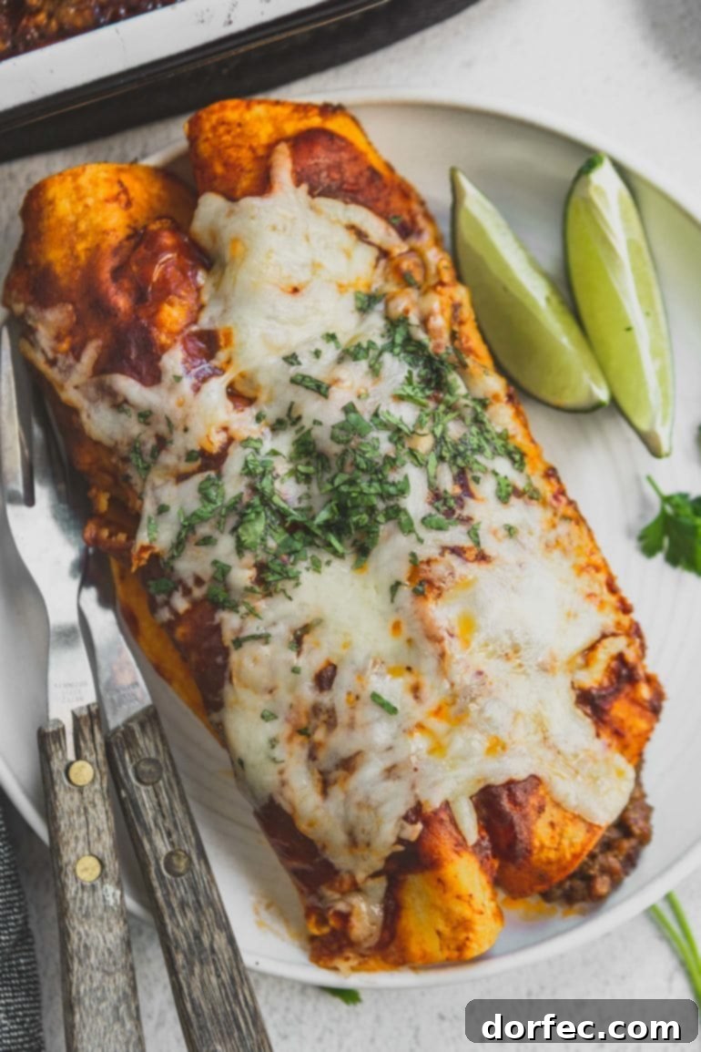 Two delicious enchiladas, covered with melted cheese and fresh chopped cilantro, served on a dinner plate with lime wedges and silverware.