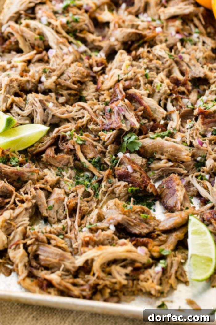 Pork carnitas, both crispy and shredded, neatly arranged on a sheet pan with fresh lime wedges and garnished with chopped cilantro.