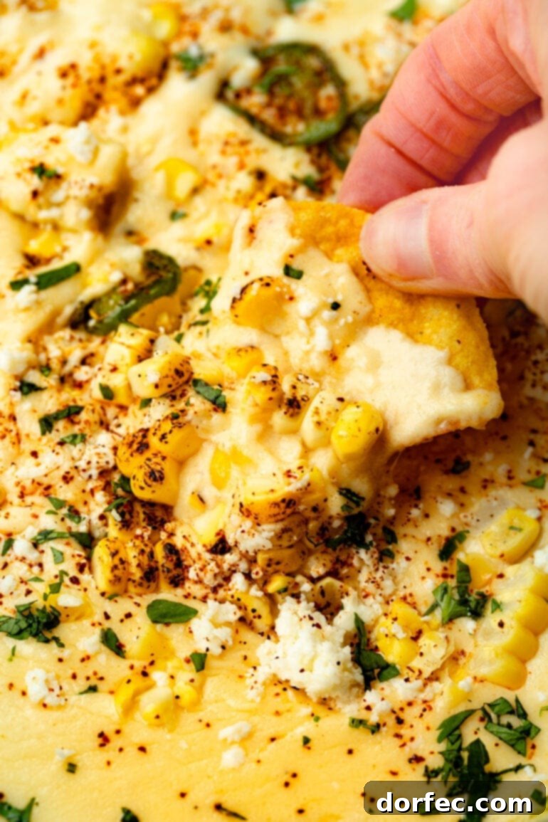 Someone lifting a corn tortilla chip from a skillet of creamy Elote Queso Dip, showcasing its enticing texture and vibrant colors.
