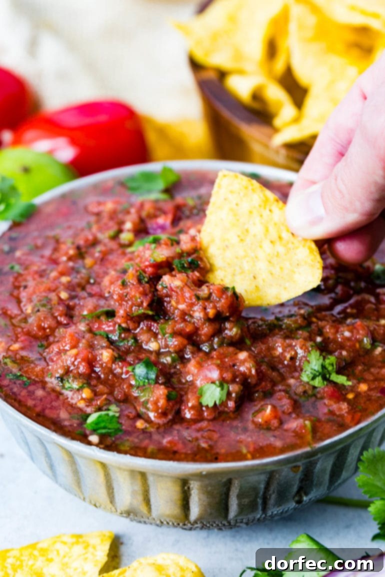 A tempting bowl of fresh homemade salsa, with a tortilla chip being dipped into it. The salsa is garnished with cilantro leaves, and in the background, there are fresh cilantro, Roma tomatoes, a lime, and a wooden bowl of chips.
