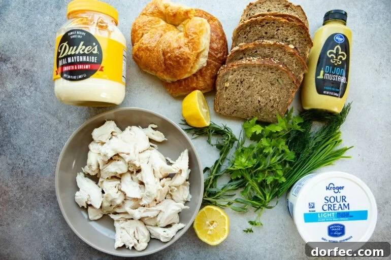 Ingredients like a jar of mayonnaise, dijon mustard, sour cream, lemon, fresh herbs, rotisserie chicken, croissants, and yummy seedy bread. 