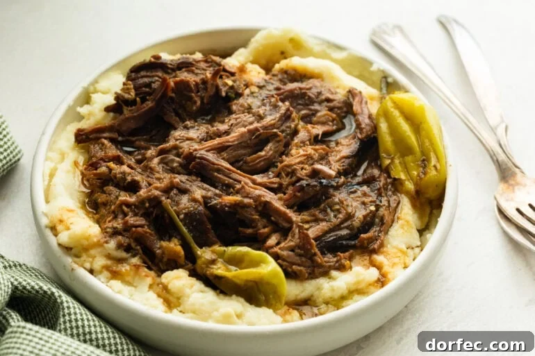 Mississippi Pot Roast Slow Cooker Recipe for Tender, Flavorful Beef 2 a large white serving bowl full of creamy mashed potatoes topped with juicy shredded pot roast with whole pepperoncinis scattered around the bowl