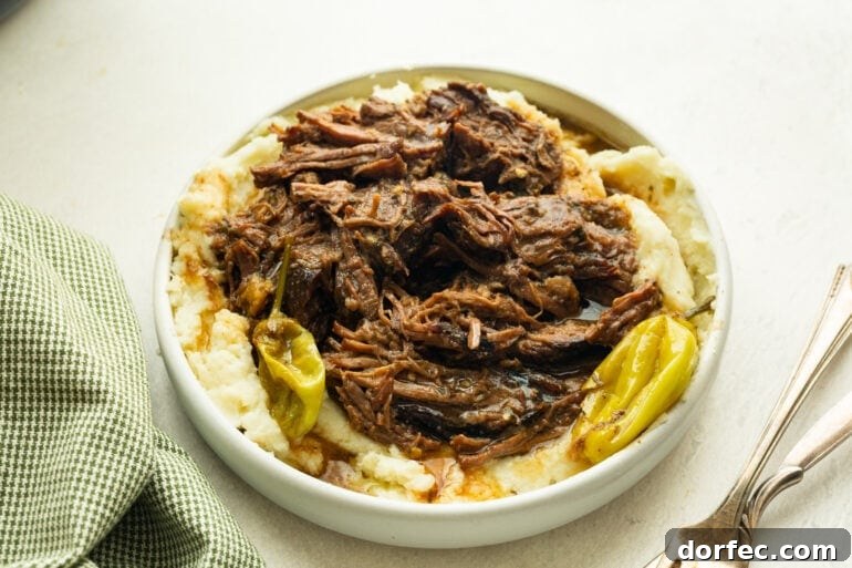 Mississippi Pot Roast Slow Cooker Recipe for Tender, Flavorful Beef 3 a large serving bowl full of creamy mashed potatoes topped with juicy, tender shredded beef with pepperoncinis scattered around it