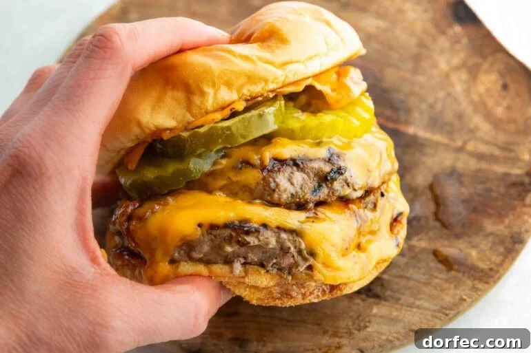 a hand holding a juicy double patty cheeseburger with dill pickle slices and a toasted bun