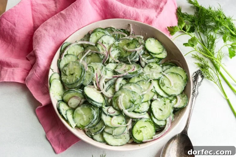 A large white bowl of creamy cucumber salad with thinly sliced English cucumbers and red onion coated in a sour cream dill dressing photographed from overhead on a light surface.