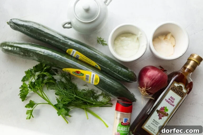 An overhead flat lay of creamy cucumber salad ingredients including whole English cucumbers, a bunch of fresh dill, a sliced red onion, and small bowls of sour cream and mayonnaise on a white marble surface.