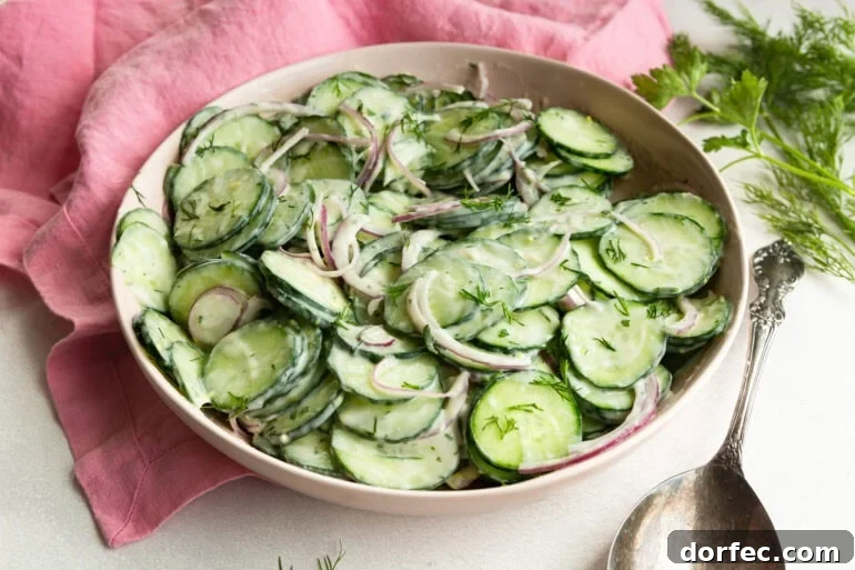 A side angle shot of a white serving bowl filled with veggies covered in a sour cream dressing sitting on a wooden surface with fresh dill and a linen napkin alongside.