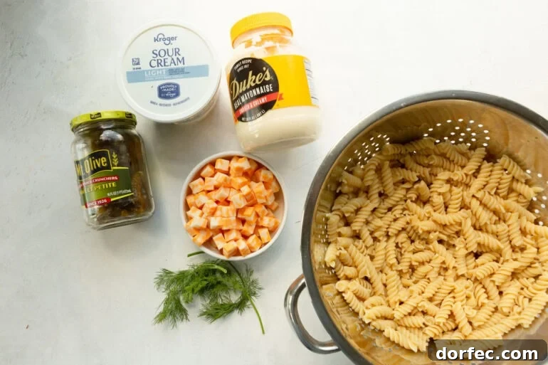 A colander filled with rotini pasta, dill, pickles, cubes of cheese, dukes mayo, and sour cream.